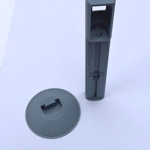 May include: A gray plastic TSHOT 10ml syringe holder with a vertical column featuring a graduated scale and the text TRT Solutions, accompanied by a separate circular base with a central rectangular slot for mounting the column.
