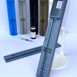 Puede incluir: Varios dispositivos médicos de plástico gris, azul, negro y marrón con el texto "TRT Solutions". También hay una jeringa y un pequeño vial. Los dispositivos tienen una escala del 1 al 10.