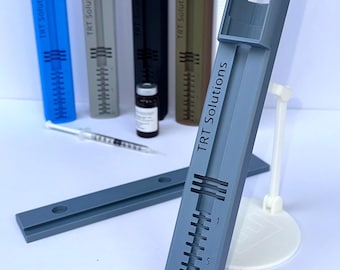 May include: A gray plastic syringe holder with the text TRT Solutions printed on the side, featuring a numbered scale from 1 to 10. Additional holders in blue, light gray, black, brown, and white are visible, along with a syringe and vial.