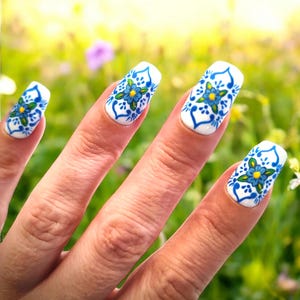 May include: Close-up of a hand with painted fingernails. Each nail is white with a blue and yellow floral design. The design features a central blue flower with yellow accents, surrounded by blue decorative patterns.