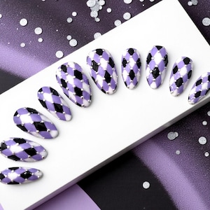 May include: A set of oval-shaped false nails with a harlequin pattern in black, white, and lavender. Each nail is adorned with small silver studs along the base. The nails are arranged on a white surface against a purple background.