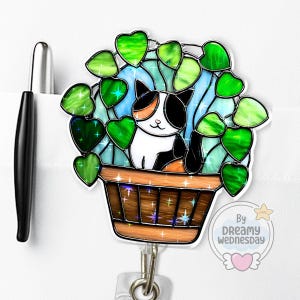 May include: A badge reel featuring a calico cat in a flower pot with green heart-shaped leaves. The pot is brown with star accents. The background is light blue. A black pen is in the background.