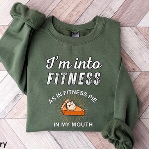 Może przedstawiać: Zielona wojskowa bluza z napisem "I'm into FITNESS as in FITNESS PIE IN MY MOUTH". Grafika zawiera kawałek ciasta z uśmiechniętą twarzą. Bluza ma długie rękawy i okrągły dekolt.