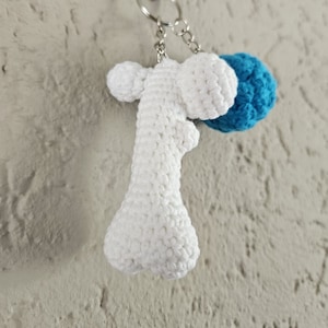 Op de afbeelding: Witte gebreide sleutelhanger in de vorm van een bot met een blauwe gebreide bal eraan bevestigd.