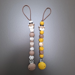 Può includere: Due clip per ciuccio per bambini con perline a forma di cervo. Una clip presenta perline marroni, bianche e beige, mentre l'altra ha perline gialle, marroni e beige. Entrambi hanno un anello marrone e una clip in basso.