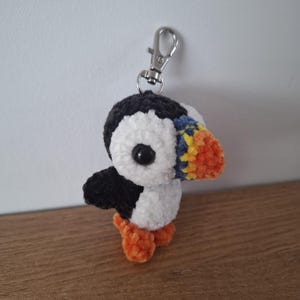 Può includere: Un portachiavi a forma di puffin fatto a crochet con un corpo bianco e nero, piedi arancioni e un becco giallo e arancione. Il portachiavi ha un moschettone in metallo argentato.