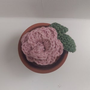 Op de afbeelding: Een gehaakte roze bloem met groene bladeren in een terracotta pot.