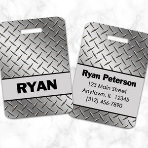 Könnte beinhalten: Zwei silberne Gepäckanhänger mit Riffelblechmuster. Auf einem steht der Name "RYAN" in fetten schwarzen Buchstaben. Der andere zeigt "Ryan Peterson" und Kontaktinformationen.