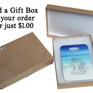 Puede incluir: Una caja de regalo marr&oacute;n con un interior blanco y una etiqueta de regalo blanca con el texto "life is BETTER at the BEACH" en letras negras. La etiqueta de regalo tiene una escena de playa azul y blanca con olas y un cielo azul.