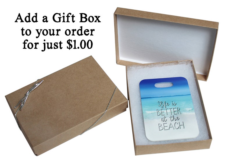 Puede incluir: Una caja de regalo marr&oacute;n con un interior blanco y una etiqueta de regalo blanca con el texto "life is BETTER at the BEACH" en letras negras. La etiqueta de regalo tiene una escena de playa azul y blanca con olas y un cielo azul.