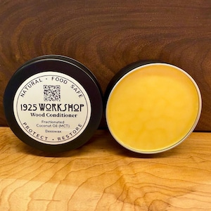 May include: A round tin container with a black lid and a label that reads "1925 WORKSHOP Wood Conditioner". The label also includes the text "Natural Food Safe", a QR code, and the words "Protect" and "Restore". The tin is open and the container is filled with a yellow, waxy substance.