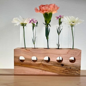 May include: A wooden flower vase with six clear glass test tubes for holding flowers. The vase is made of light brown wood with a natural grain pattern. The vase is filled with pink, white, and light purple flowers.