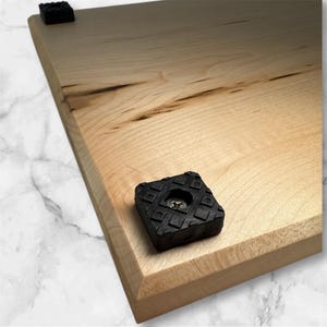 May include: A light-colored wooden cutting board with a beveled edge. The board has dark wood grain patterns and is fitted with four black rubber feet, each approximately 1 inch square, to prevent slipping.