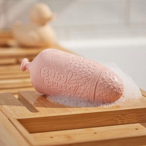 May include: Pink bath toy shaped like a bottle with the words "Baby Squeezy" on the side. The toy is made of textured silicone and has a small spout at the top.
