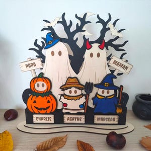 Peut inclure: Une décoration d'Halloween en bois représentant deux grands fantômes étiquetés Papa et Maman, et trois petits fantômes déguisés en citrouille, épouvantail et sorcière, étiquetés Charlie, Agathe et Marceau, devant un arbre noir.