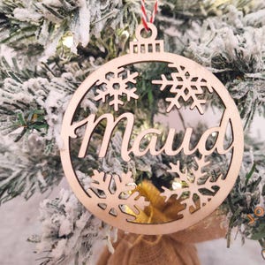 Peut inclure: Ornement de Noël en bois avec le nom "Maud" découpé, entouré de flocons de neige. L'ornement est suspendu à une ficelle rouge et est placé sur un sapin de Noël enneigé, créant une scène festive.