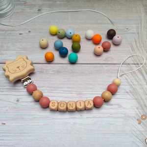Peut inclure: Un hochet en bois pour bébé avec un motif de tête de lion et un clip. Le hochet est composé d'une chaîne de perles en bois avec des perles en silicone rose et des perles en bois en forme de lettres qui épellent le nom "SONIA".