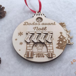 Peut inclure: Ornement de Noël en bois avec un ruban rouge, représentant une cheminée avec des chaussettes marquées "Papa", "Mael", "Lena" et "Maman". L'ornement porte l'inscription "20 Dodos avant Noël".
