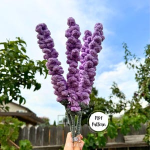 Könnte beinhalten: Ein gehäkelter Lavendelblumenstrauß in Violetttönen. Der Strauß wird in einer Hand gehalten und der Text "PDF Pattern" ist in einem Kreis sichtbar.