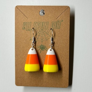 May include: A pair of Halloween-themed earrings shaped like candy corn. Each earring features a white top, an orange middle, and a yellow bottom. The earrings hang from silver-colored hooks and are displayed on a brown card with the text "ALL SPUN OUT".