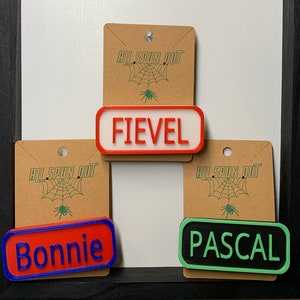 Könnte beinhalten: Drei individuell gestaltete Namensschilder mit einem Spinnennetz-Design. Die Namensschilder sind rot, blau und grün mit weißen Buchstaben. Die Namen auf den Schildern sind "Fievel", "Bonnie" und "Pascal".