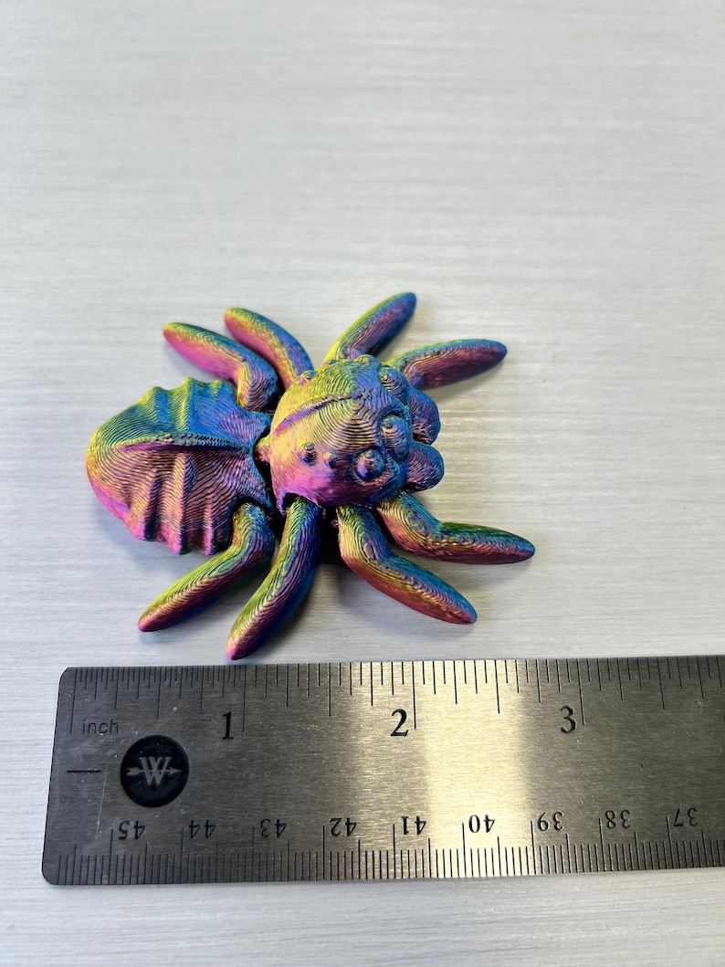 May include: A iridescent rainbow colored 3D printed spider figurine measuring approximately 1 inch in length. The spider is sitting on a ruler.
