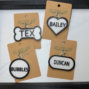 May include: Four personalised dog tags with black outlines and white lettering. The tags are shaped like a bone, a heart, a circle, and an oval. The names on the tags are Tex, Bailey, Bubbles, and Duncan.