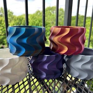 May include: Five wavy-textured ceramic planters in different colors: white, blue, purple, gray, and red. The planters are arranged in a circle on a metal surface.