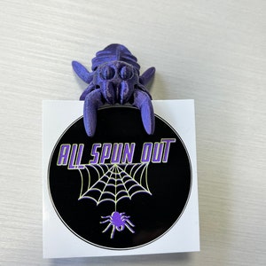 May include: Purple 3D printed spider figurine holding a black and white sticker with the text "All Spun Out" and a spider web design.