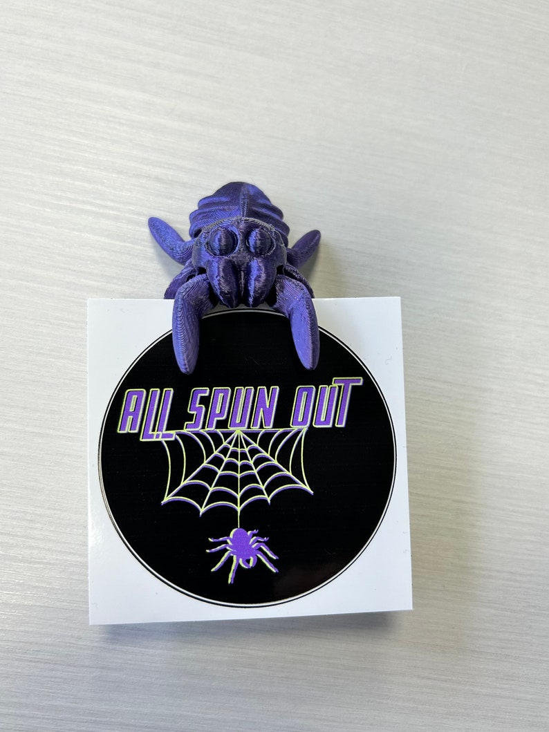 May include: Purple 3D printed spider figurine holding a black and white sticker with the text "All Spun Out" and a spider web design.