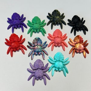Puede incluir: Una colección de diez objetos decorativos con forma de araña de colores. Las arañas son de varios colores, incluyendo morado, verde, rojo, rosa y negro. Cada araña tiene ocho patas y un cuerpo detallado. Los objetos están dispuestos sobre una superficie blanca.