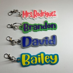May include: Four colorful 3D printed keychains with names on them. The keychains are attached to silver key rings. The names are "Mrs. Rodriguez", "Brandon", "David", and "Bailey".