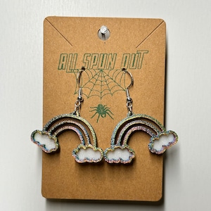 May include: A pair of rainbow and cloud earrings with silver hooks. The earrings are made of glitter and have a holographic finish. The earrings are on a brown card with the text "All Spun Out" printed on it.