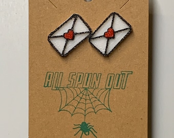 May include: A pair of white envelope earrings with red hearts, displayed on a brown card with the text "All Spun Out" and a green spider web with a spider.