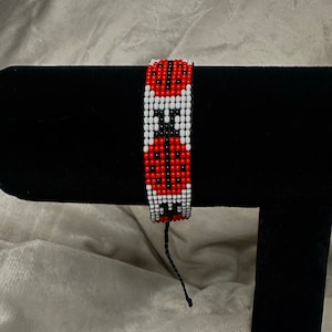 May include: A red, white and black beaded bracelet with a geometric pattern. The bracelet is tied with a black string.