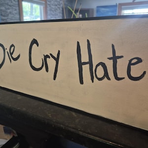 May include: A rectangular sign with the words "Die Cry Hate" in black handwritten lettering on a cream-colored background. The sign has a black border and is displayed on a dark surface.