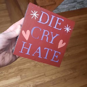 May include: A red wooden sign with the words "*DIE* CRY HATE" in purple lettering and two pink hearts.