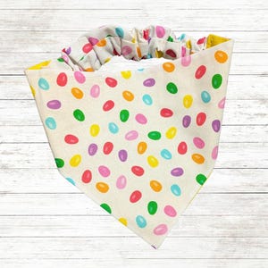 May include: A white bandana with a colorful jelly bean pattern. The bandana has a ruffled edge and is tied around the neck.