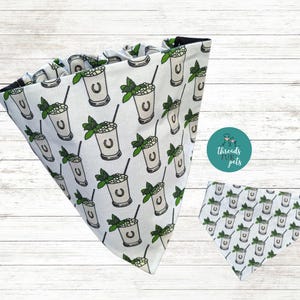 May include: White dog bandana with a repeating pattern of mint julep cocktails. Each cocktail illustration includes a horseshoe, mint sprig, and a black straw. The bandana is triangular and has a black border.