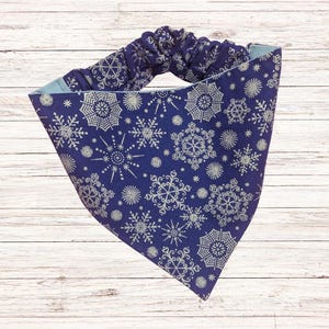 May include: A navy blue headband with a snowflake pattern. The fabric features a repeating design of white snowflakes of various sizes. The headband has a gathered, elasticated section for a comfortable fit and a light blue lining.
