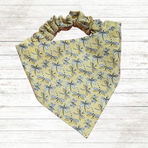 May include: Yellow dog bandana with a repeating dragonfly pattern in blue, gray, and yellow. The bandana has an elastic band for easy wear. The fabric is a light yellow color.