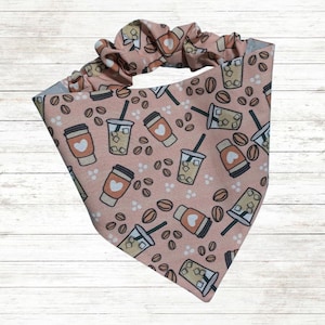May include: A pink dog bandana with a coffee theme. The bandana features a repeating pattern of coffee cups, iced coffee, and coffee beans. The top of the bandana has a matching pink scrunchie.