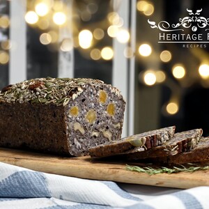 May include: A loaf of seeded bread with visible nuts and dried fruit, partially sliced, on a wooden cutting board. The bread is dark brown with a variety of seeds on top. The image includes the text "HERITAGE FOOD RECIPES".