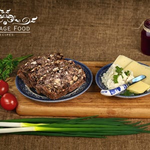May include: A wooden cutting board with a blue and white patterned plate holding slices of dark bread. A second blue and white patterned plate holds cheese and a small knife. A jar of red jam, a bunch of green onions, and red tomatoes are also on the board.