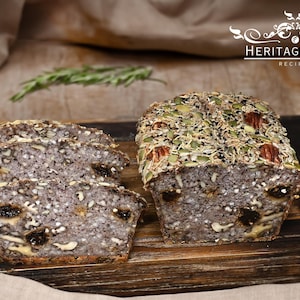 May include: Two loaves of seeded bread, one sliced, on a wooden board. The bread is dark with visible seeds, nuts, and dried fruit. The top is covered with pumpkin seeds and pecans. A sprig of rosemary is in the background. The text "HERITAGE FOOD RECIPES" is in the upper right.