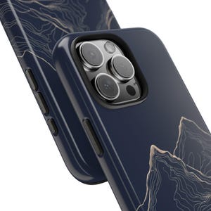 May include: A navy blue phone case with a gold abstract mountain design. The case has a raised camera bump with three lenses.