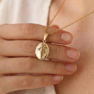 May include: A gold-colored oval locket necklace with a delicate floral design. The pendant hangs from a fine chain. The locket is held by a person wearing a white shirt.