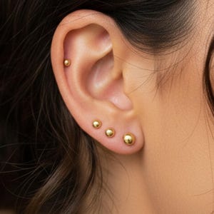 May include: Close-up of an ear with four gold ball stud earrings. The earrings are of varying sizes, with the largest at the bottom and the smallest at the top. The ear is set against a blurred background.