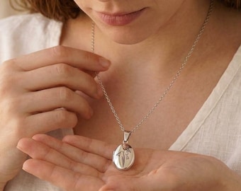 May include: Silver oval pendant necklace with a delicate floral design. The pendant is suspended from a fine chain. The necklace is held in a person's hand, showcasing the pendant's details. The background is a soft, neutral color.