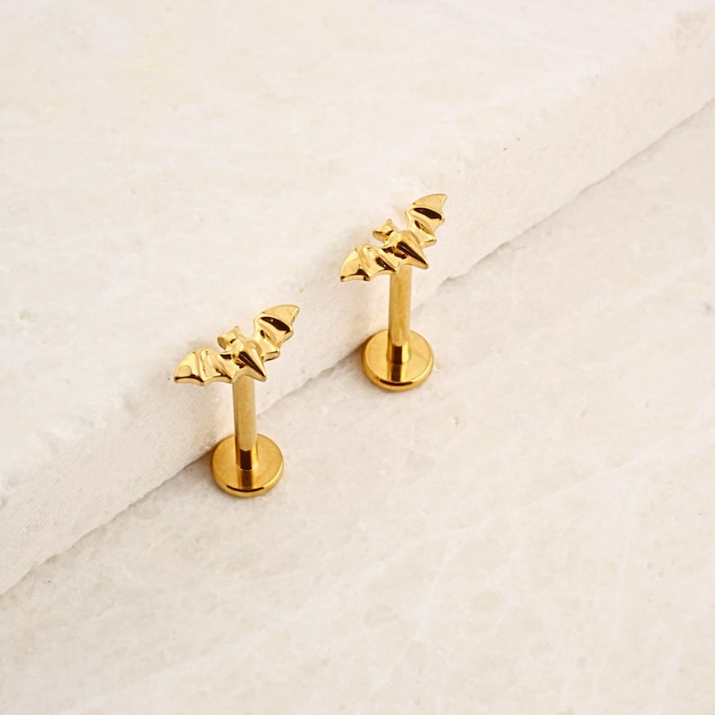 K&ouml;nnte beinhalten: Zwei goldfarbene Lippenringe in Form einer Fledermaus. Die Ringe sind aus Chirurgenstahl und haben eine flache R&uuml;ckseite.